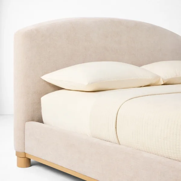 Arched headboard detail of an upholstered teakwood bed with textured beige upholstery