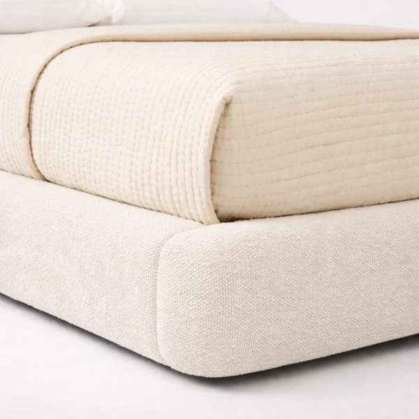 Padded frame detail of a fully upholstered bed with soft corners and textured neutral fabric